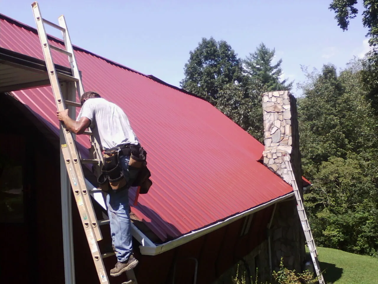 Roofer on a ladder beside a red metal roof and stone chimney — Soffit & Fascia Repair by Roofing Brothers in Lefor